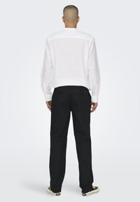 Uomo in piedi di spalle che indossa una camicia bianca a maniche lunghe, pantaloni neri e sneaker bianco e nere su uno sfondo chiaro e uniforme.