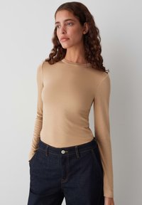 Lange mouwen, aansluitend top in beige, gemaakt van een gladde stof. Gecombineerd met donkere denim broek met een knoop en oranje stikdetails.