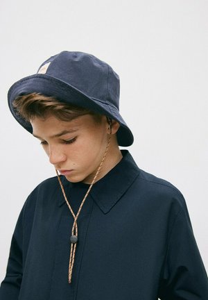 Niño con un sombrero tipo pescador azul marino con correa para la barbilla y camiseta azul marino a juego, mirando hacia abajo sobre un fondo claro y liso.