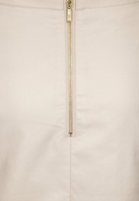 Rundad kjol i mjukt beige tyg. Har en guldfärgad dragkedja längs mitten, med synliga sömmar och en slät yta. Enkelt och minimalistiskt design.