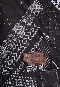 Tissu noir avec des motifs géométriques blancs et une pièce en cuir marron portant l'étiquette "BRUNOTTI", cousue près d'une couture.