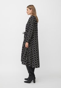Schwarzes Kleid mit einem gepunkteten Muster in Creme, langen Ärmeln und einem taillierten Gürtel, kombiniert mit knielangen schwarzen High Heels Stiefeln.