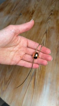 Fém ovális medál egy kézben, vékony fonott láncra fűzve. A medál tükröződő felülettel rendelkezik, amely gazdag barna árnyalatot mutat.