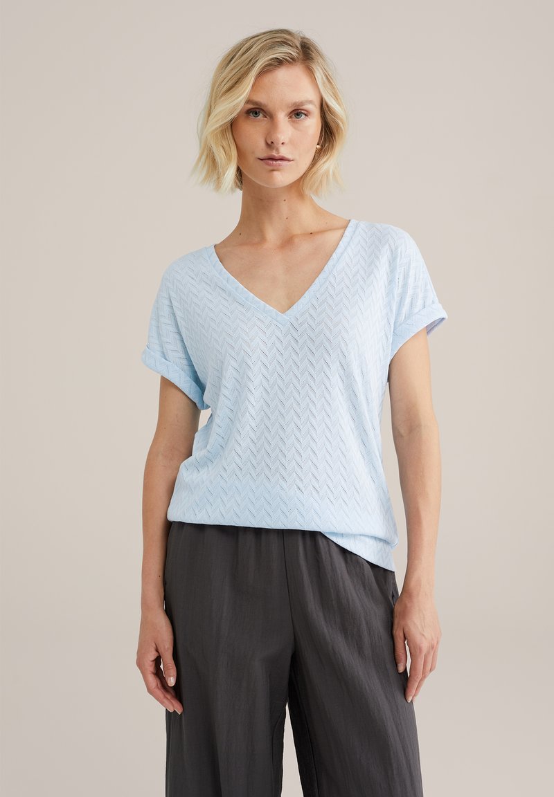 Gaiši zilas V-veida krekls ar teksturētu adījumu, ar rolled piedurknēm un brīvu piegriezumu, savienots ar tumši pelēkiem plašiem biksēm.