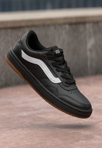 Black leather sneaker with white stripes, textured upper, and lace closure. Brown rubber sole for traction. Classic low-top design.