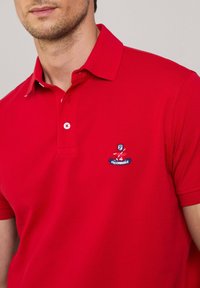 Maglietta polo rossa in tessuto testurizzato con colletto classico. Presenta una chiusura con bottoni bianchi e logo ricamato sul petto.