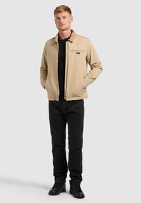 Jeune homme debout les mains dans les poches, portant une veste beige, une chemise noire, un pantalon noir et des chaussures noires sur un fond uni.