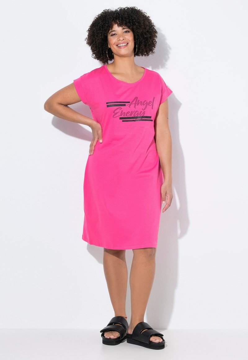 Rosa klänning med korta ärmar som har texten "Angel Energy" i svart glitter, med en avslappnad passform och knä-lång nederdel, tillsammans med svarta sandaler.