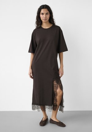 Kvinde stående iført en løs mørk midi-kjole med korte ærmer, sideslids, blonderkant og matchende flade sko mod en ensfarvet baggrund.