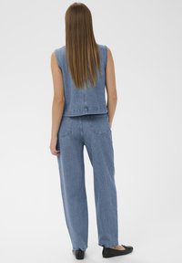 Frau mit langen, glatten Haaren, die eine ärmellose Jeansweste, passende weite Jeans und schwarze Ballerinas trägt, steht vor einem einfarbigen Hintergrund.