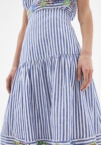 Sininen ja valkoinen pystylinjainen mekko, jossa on kukkakirjailua helmassa, yllään henkilöllä, jolla on näkyvä käsivarttattoo.