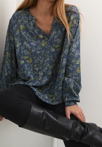 Blouse florale bleu foncé et vert avec une coupe décontractée. Présente un col en V, des manches longues et un motif de tissu texturé.