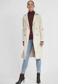 Beige trenchcoat med dobbeltradet design, stor krage og mørke knapper; brukt over en burgunderrød genser med høy hals, kombinert med blå jeans og brune ankelstøvler.