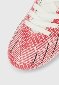 Αθλητικό παπούτσι με κόκκινο και λευκό μοτίβο, λευκά κορδόνια και μαύρες σχισμές στην περιοχή των δακτύλων, εμφανιζόμενο σε λευκό φόντο.