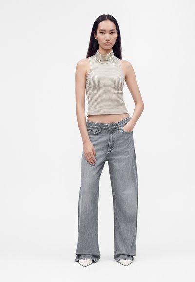 Jeune femme aux longs cheveux lisses porte un haut à col roulé rayé sans manches, un jean évasé délavé et des chaussures blanches pointues, debout la main dans la poche.
