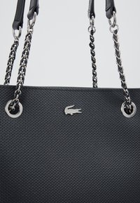 Sac fourre-tout noir texturé avec des poignées en chaîne argentée et des accents métalliques, présentant un logo argenté à l'avant.