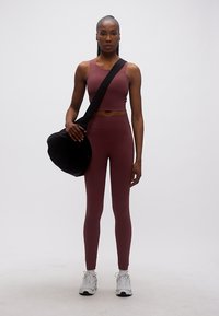 Γυμνασμένη γυναίκα με συνδυαστικό σετ γυμναστικής σε μπορντό χρώμα και λευκά αθλητικά παπούτσια στέκεται κρατώντας μια μαύρη τσάντα γυμναστηρίου στον ώμο της.