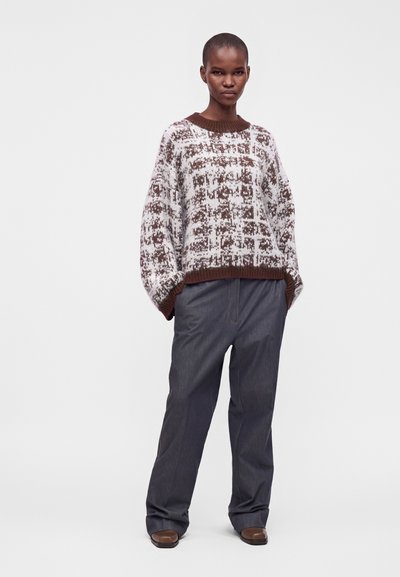 Personne debout portant un pull ample marron et blanc à motifs, un pantalon large foncé et des chaussures marron sur un fond clair uni.