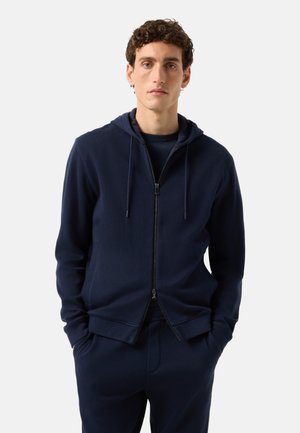 Jeune homme aux cheveux bouclés portant un sweat à capuche bleu marine zippé et un pantalon assorti, les mains dans les poches, debout devant un fond uni.