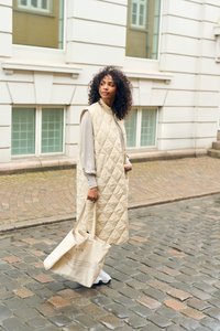 Gilet long matelassé beige avec un col haut, porté sur un haut gris clair, associé à un sac fourre-tout crème et des baskets blanches sur une rue pavée.