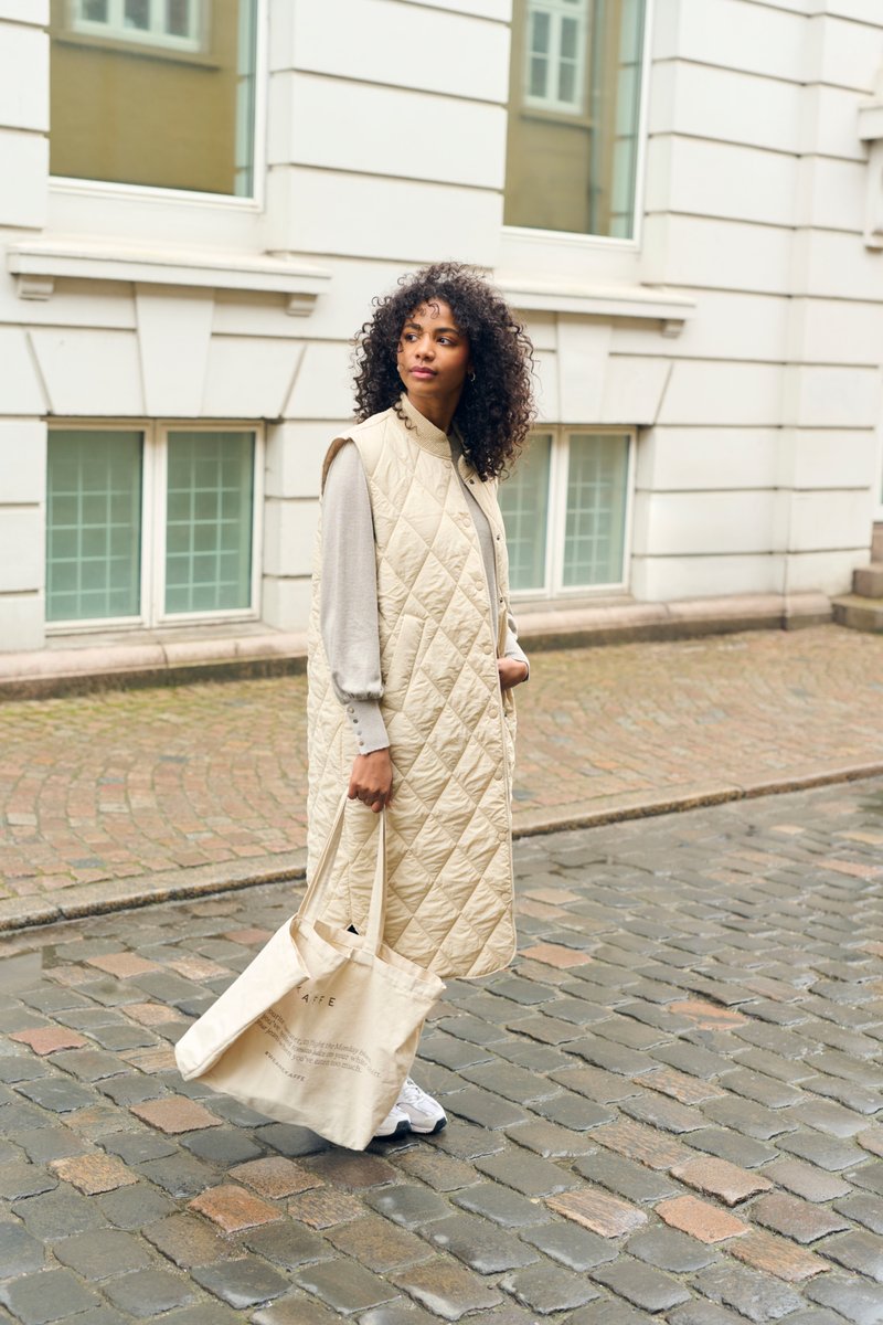 Gilet long matelassé beige avec un col haut, porté sur un haut gris clair, associé à un sac fourre-tout crème et des baskets blanches sur une rue pavée.