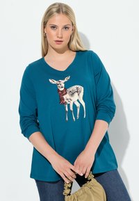 Türkises langärmliges T-Shirt mit V-Ausschnitt, auf dem ein grafisches Bild eines Rehs mit einem roten Schal zu sehen ist, kombiniert mit dunkelblauen Jeans und einer beigen Handtasche.