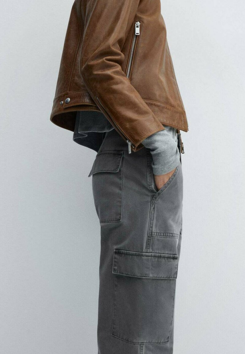 Brązowa skórzana kurtka z zapięciem na zamek, zestawiona z szarymi spodniami cargo z wieloma kieszeniami i teksturowanym wykończeniem. Pod spodem szara bluza z długim rękawem.