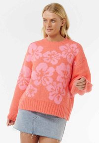 Blonde Frau, die einen flauschigen orangefarbenen Pullover mit pinken Blumenmustern und einen hellblauen Denim-Minirock vor einem weißen Hintergrund trägt.