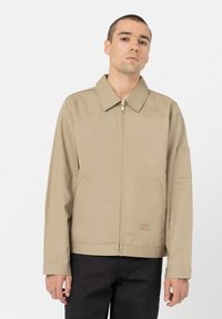 Beige zip-jakke med spiss krage, sidelommer og et lite logo-patch. Har en rett passform og jevn tekstur.