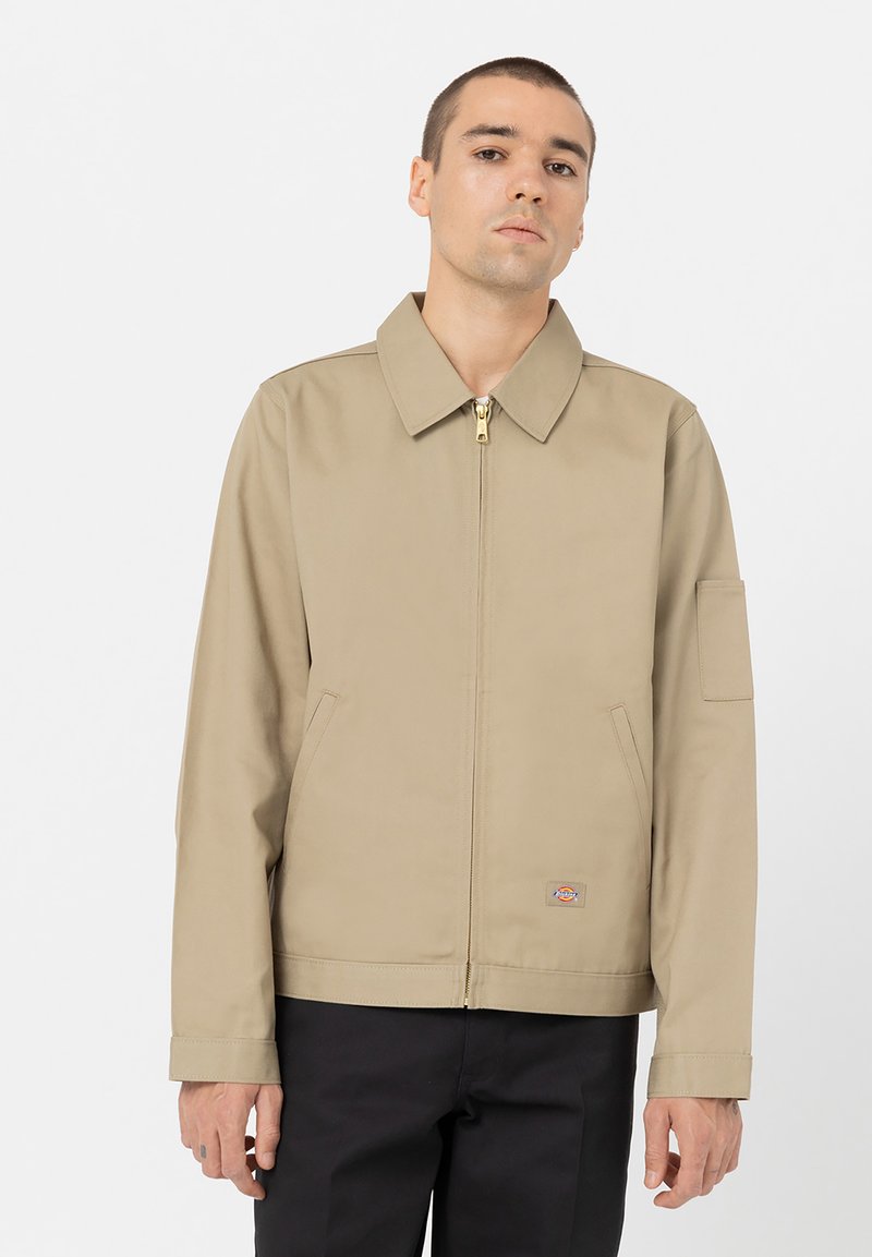 Beige zip-jakke med spiss krage, sidelommer og et lite logo-patch. Har en rett passform og jevn tekstur.
