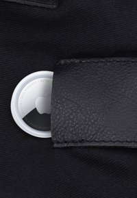 Apple AirTag teilweise in einen schwarzen Lederband eingesetzt, der an dunklem Stoff befestigt ist.