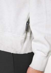 Biała bluza z teksturowanym wykończeniem, z surowym dołem i ściągaczami na rękawach, z czarną metką na bocznej szwie.