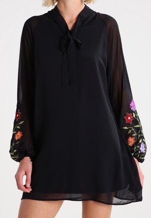 Mujer vestida con un vestido negro de mangas largas transparentes que presenta un bordado floral colorido y un escote con lazo.