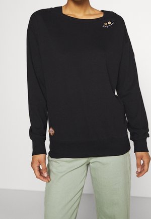 Personne portant un sweat-shirt noir à manches longues avec de petits boutons et une broderie sur le col, assorti à un pantalon vert clair.