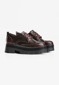 Un paio di scarpe brogue in pelle marrone scuro con suole in piattaforma nera spessa e lacci marroni abbinati, su uno sfondo bianco.