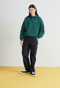 Groene pullover sweatshirt met een kraag en logo, gecombineerd met zwarte cargobroeken. Zwarte sneakers met witte veters maken de outfit compleet.