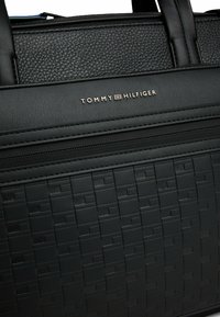 Schwarze Texturtasche mit einem gewebten Muster, glatten Akzenten und goldenem "TOMMY HILFIGER"-Logo auf der Vorderseite. Zwei stabile Griffe.