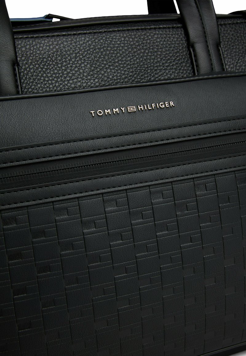 Black textured tote bag featuring a woven pattern, smooth accents, and gold "TOMMY HILFIGER" logo on the front. Two sturdy handles.