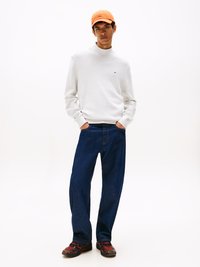 Weißer Pullover mit hohem Rippkragen, dunkelblaue, lockere Jeans und eine orangefarbene Cap. Die Sneaker haben rote Akzente und schwarze Sohlen.