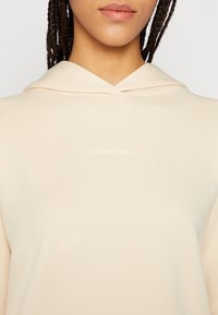 Sudadera con capucha beige de textura suave. Presenta un pequeño logo blanco de "Calvin Klein" en el frente y un diseño de bolsillo canguro.