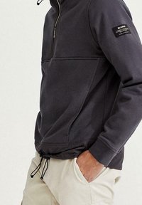 Sudadera gris con cremallera hecha de tela suave, con cuello alto, bolsillo tipo canguro y cordón ajustable en la cintura. Parche de acento negro.