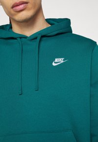 Teal mikina vyrobená z bavlněné směsi, s přední kapsou, kapucí se stahovací šňůrkou a bílým logem Nike na hrudi. Měkká textura.