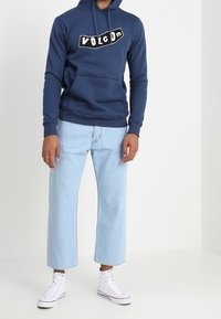 Sudadera azul marino con el logo blanco de "VULCOM", bolsillo frontal, combinada con jeans de pierna ancha de color azul claro y zapatillas altas blancas.