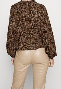 Schwarze Bluse mit kleinem braunem Blumenmuster, langen Ärmeln und lockerer Passform. Kombiniert mit beigen Lederhosen.