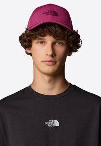 Magenta keps med broderad logotyp, kombinerad med en mörkgrå t-shirt med rund hals som har en liten vit logotyp på bröstet.