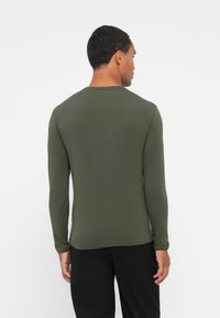 Bărbat îmbrăcat cu o cămașă lungă de culoare verde măslinie și pantaloni negri, văzut din spate, pe un fundal alb simplu.