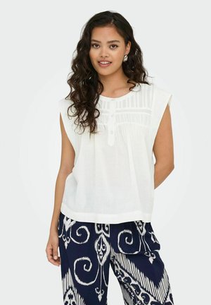 Blouse sans manches blanche avec un col rond, présentant un panneau texturé et trois boutons. Associée à un pantalon marine à motifs.