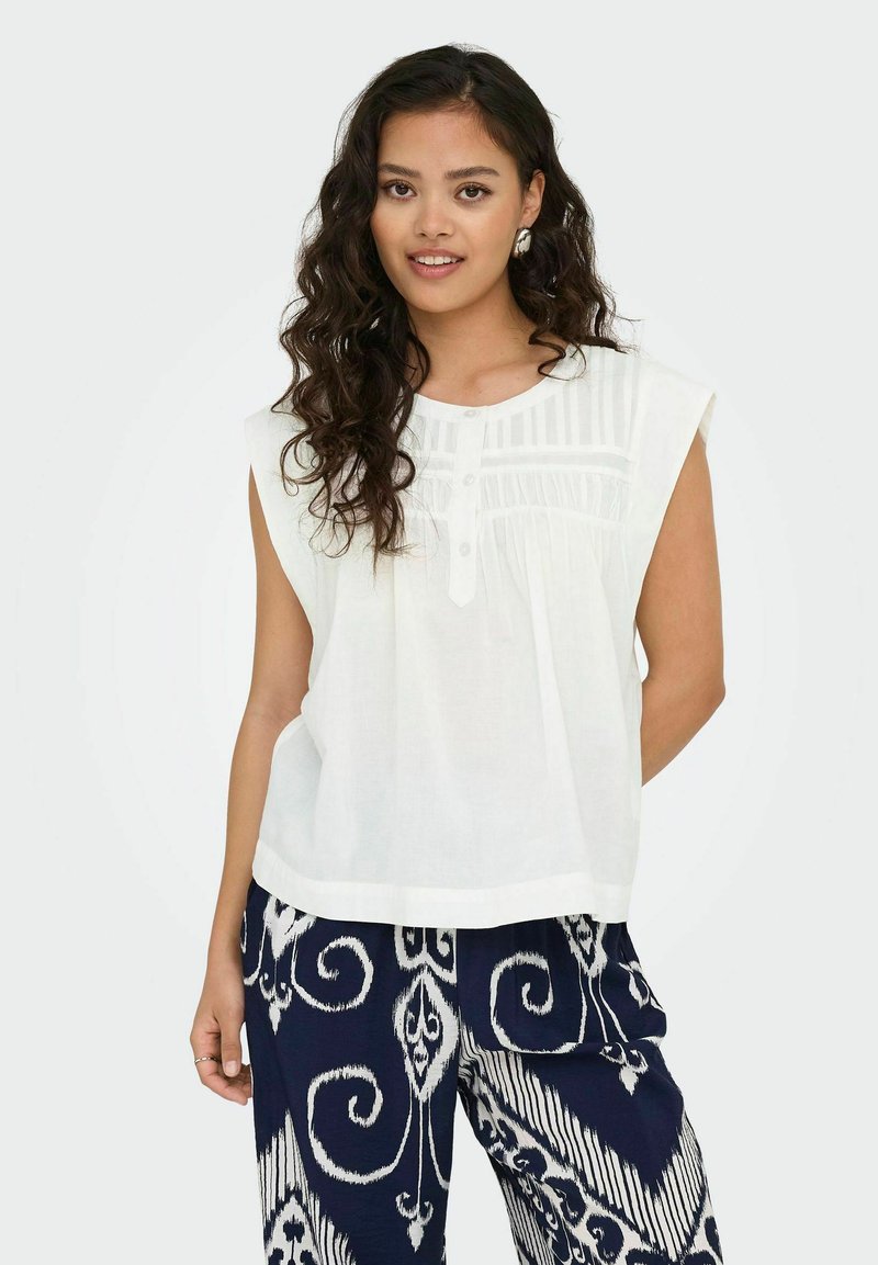Blouse sans manches blanche avec un col rond, présentant un panneau texturé et trois boutons. Associée à un pantalon marine à motifs.