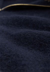 Tessuto in lana blu navy con una texture morbida, caratterizzato da sottili variazioni superficiali e un accento metallico oro riflettente sulla cerniera.