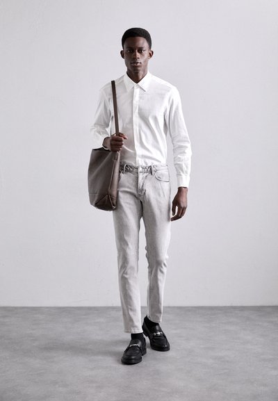 White button-up shirt, light gray jeans, and black loafers. Brown shoulder bag with textured material. Standing against a neutral background.